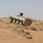Armored vehicle navigating through a sandy desert terrain with dry grass patches.