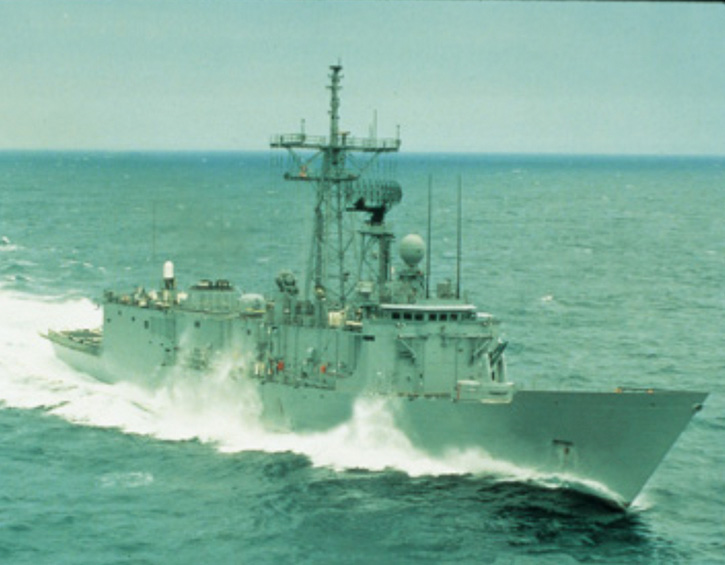 A naval warship cruising through the ocean with waves splashing against its hull.