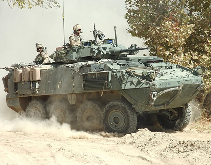 Military personnel in an armored vehicle driving through a dusty terrain with trees in the background.
