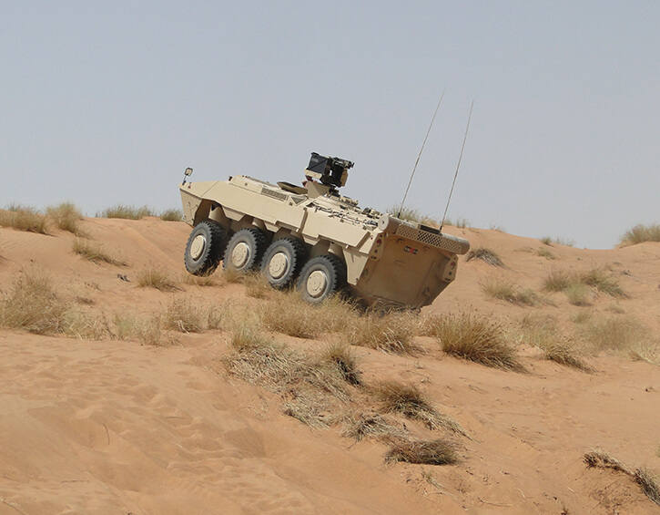 Armored vehicle navigating through a sandy desert terrain with dry grass patches.