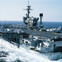 Aircraft carrier sailing with jets on deck in open sea.