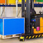 Forklift in a warehouse lifting a blue pallet box with colorful shelves in the background.