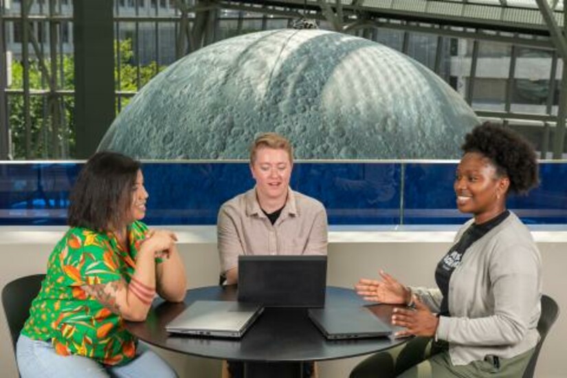Three colleagues discuss a project at a table with laptops, with a large moon model in the background.