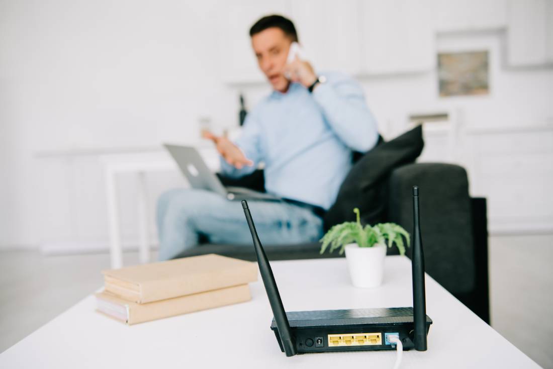 Man on phone sitting with an exasperated expression, looking at table with wifi router, books and small plant