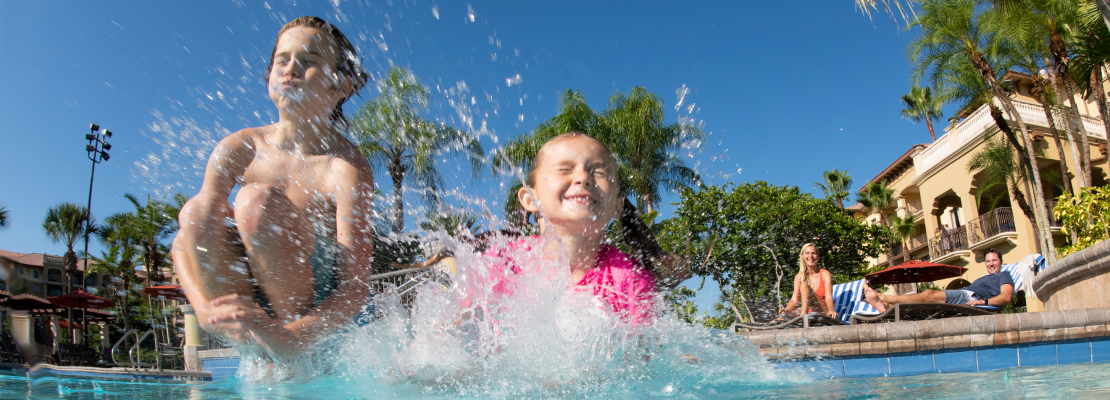 Two children cannonballing into a pool while their parents watch from poolside lawn chairs.