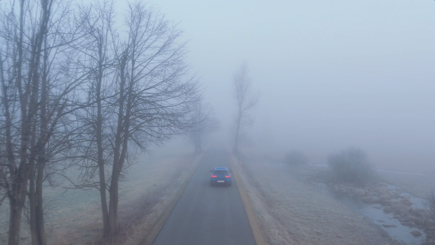 A blue car driving along a rural road surrounded by fog