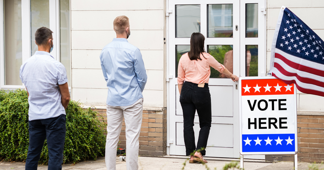 Invidividuals lined up outside, with a vote here sign beside the door