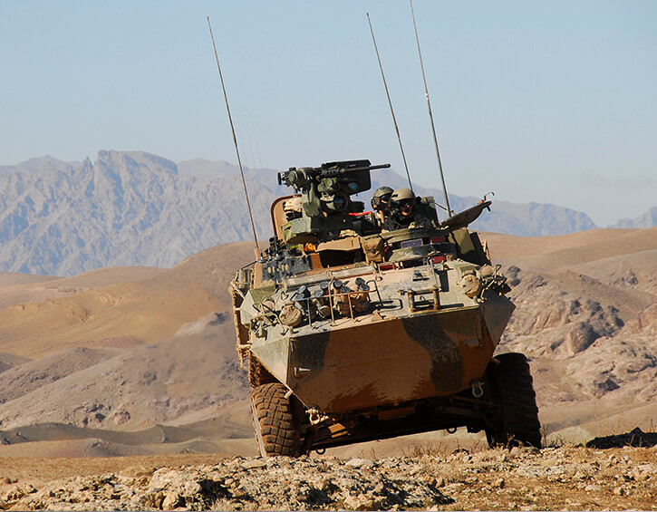 Armored military vehicle traversing rugged terrain with mountains in the background.