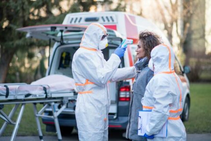 Healthcare workers in protective gear check a woman's temperature near an ambulance.