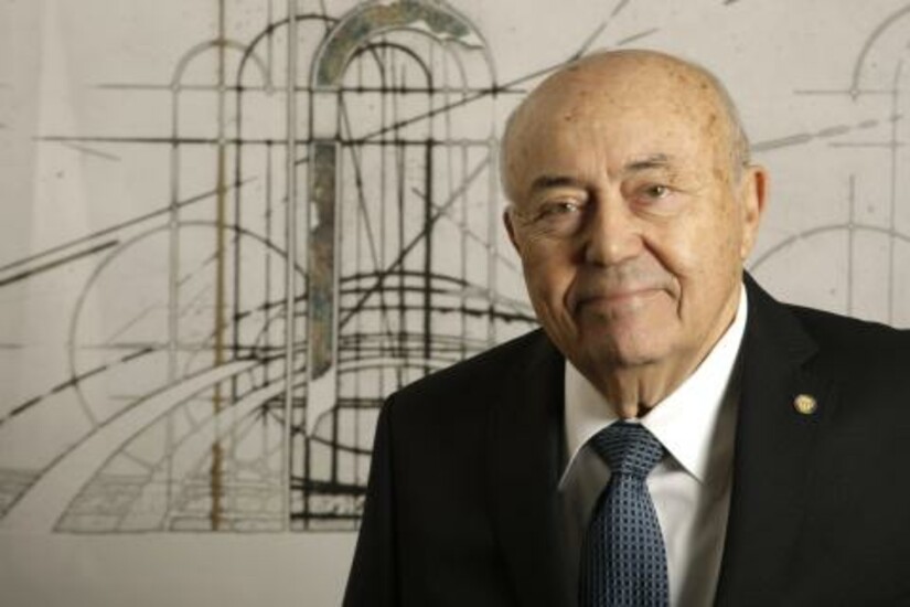 Andrew Viterbi in a suit and tie, standing in front of an abstract architectural background.