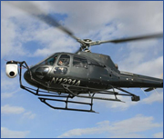 A black helicopter with a camera mounted on the front flying against a blue sky with clouds.