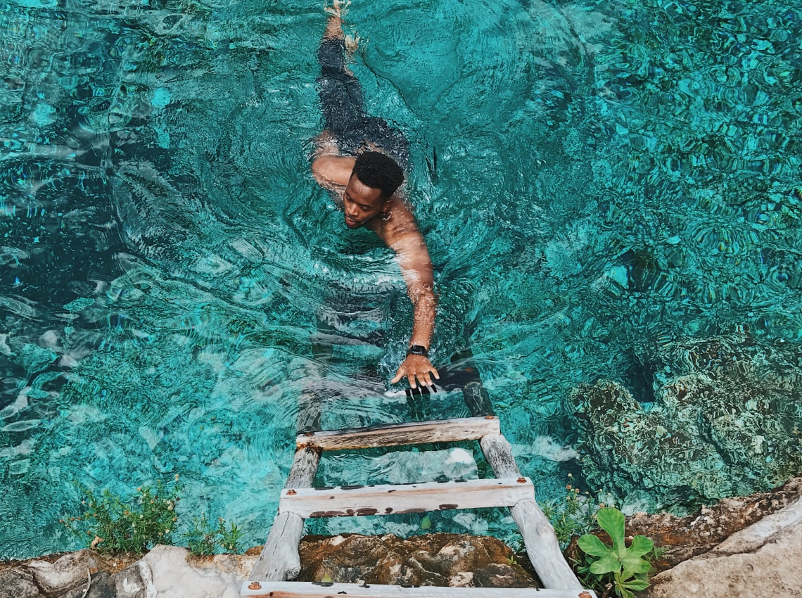 A man swimming towards a wooden ladder in crystal clear turquoise water.