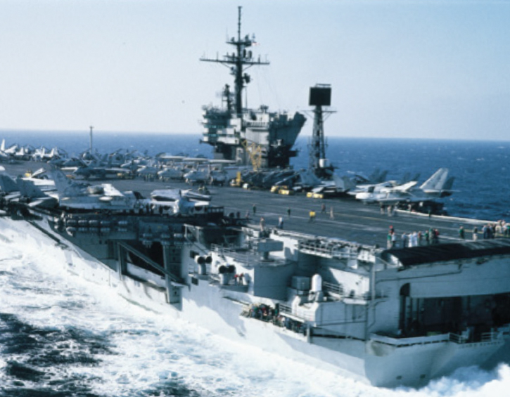 Aircraft carrier sailing with jets on deck in open sea.