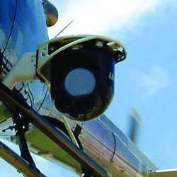 Close-up of a surveillance camera mounted on a helicopter against a clear blue sky.