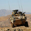 Armored military vehicle traversing rugged terrain with mountains in the background.