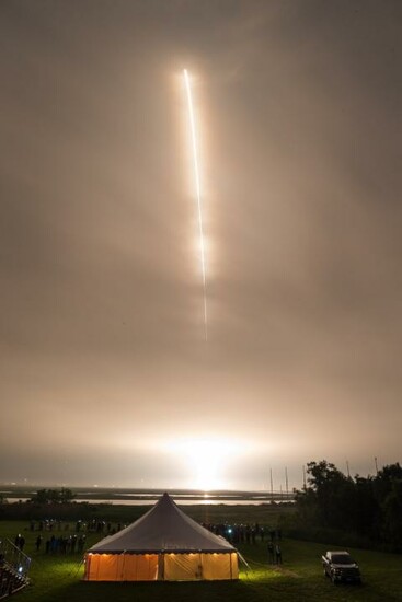 A rocket launches into the night sky, leaving a bright trail above a tent and spectators below.