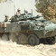 Military personnel in an armored vehicle driving through a dusty terrain with trees in the background.
