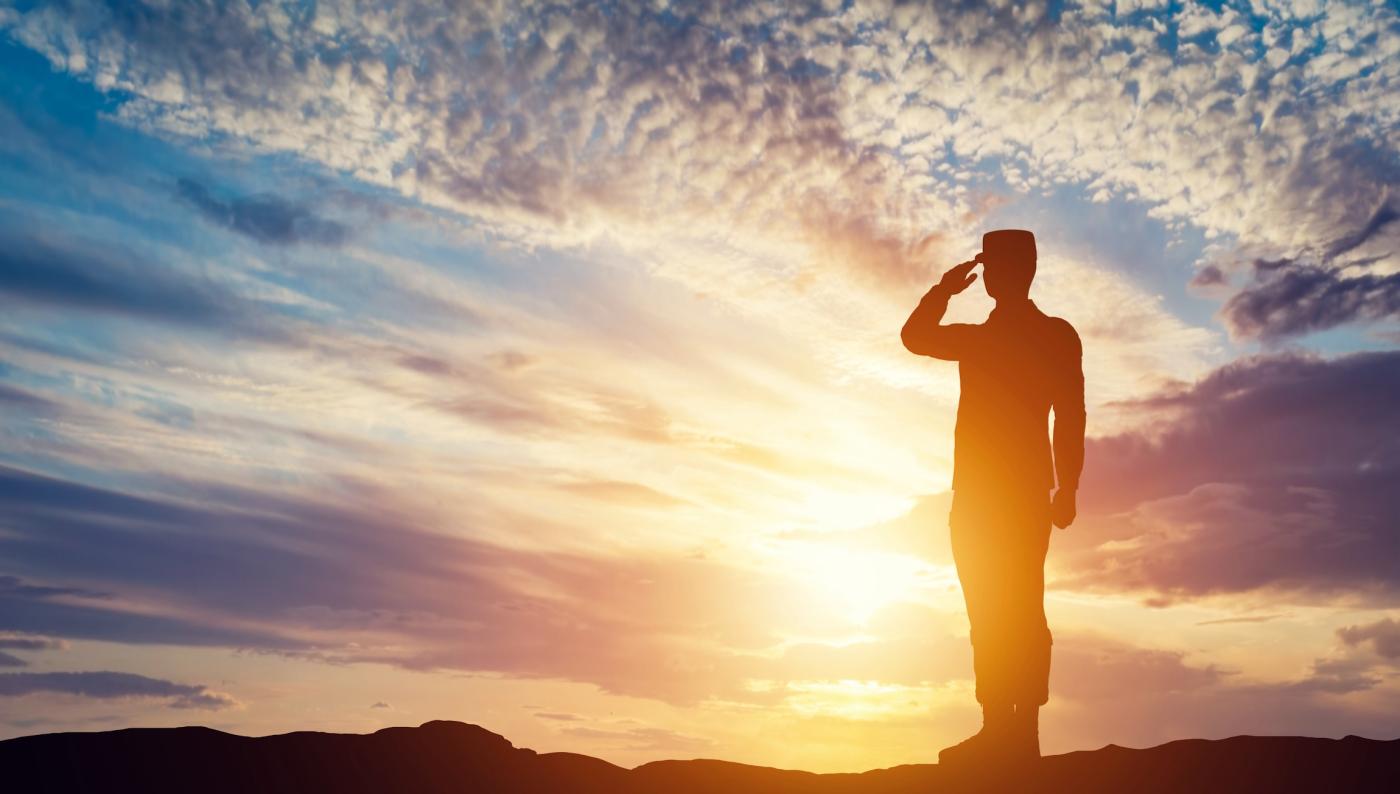 A service member saluting in provile against a yellow, purple and blue sunset